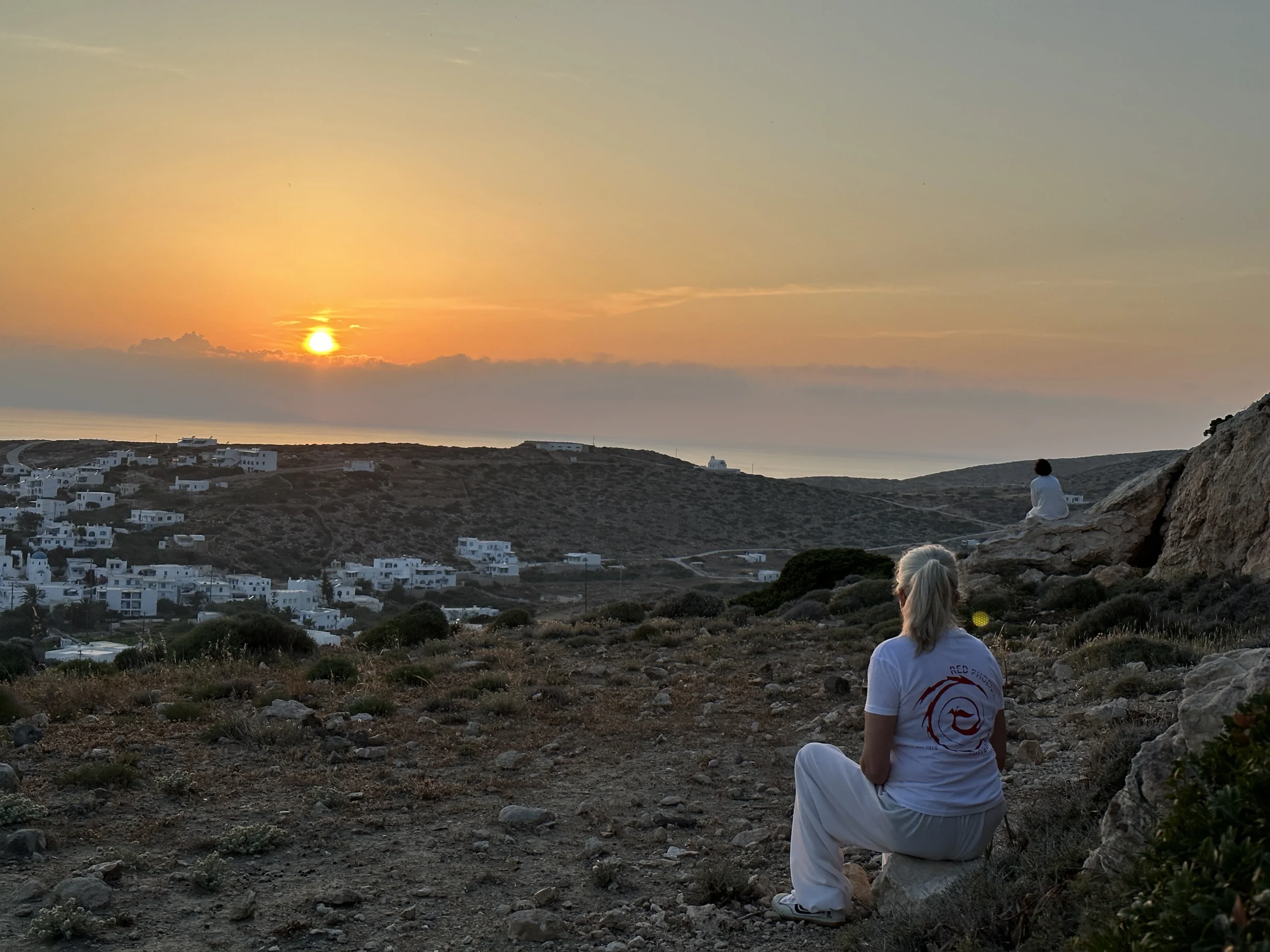 Zwei Personen meditieren auf einem Berg mit Blick aufs Meer beim Sonnenuntergang.